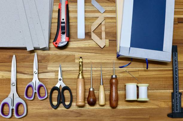 A selection of bookbinding tools, including scissors, a cutter, thread, and various specialized instruments on a wooden surface.