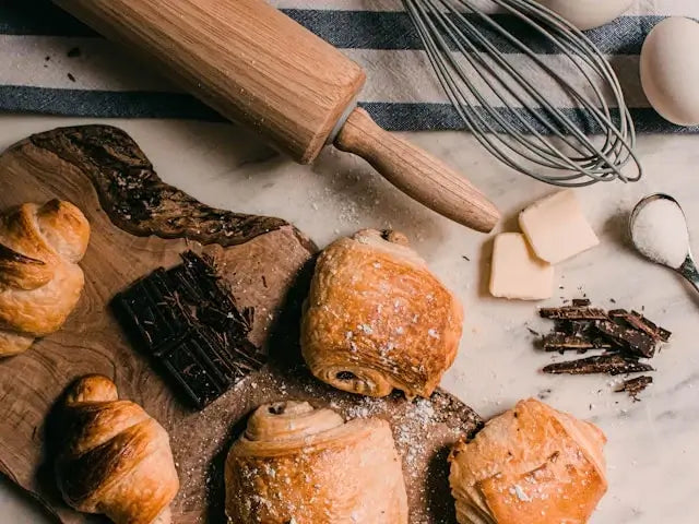 Freshly baked croissants, chocolate, butter, and eggs with a rolling pin and whisk on a marble countertop.